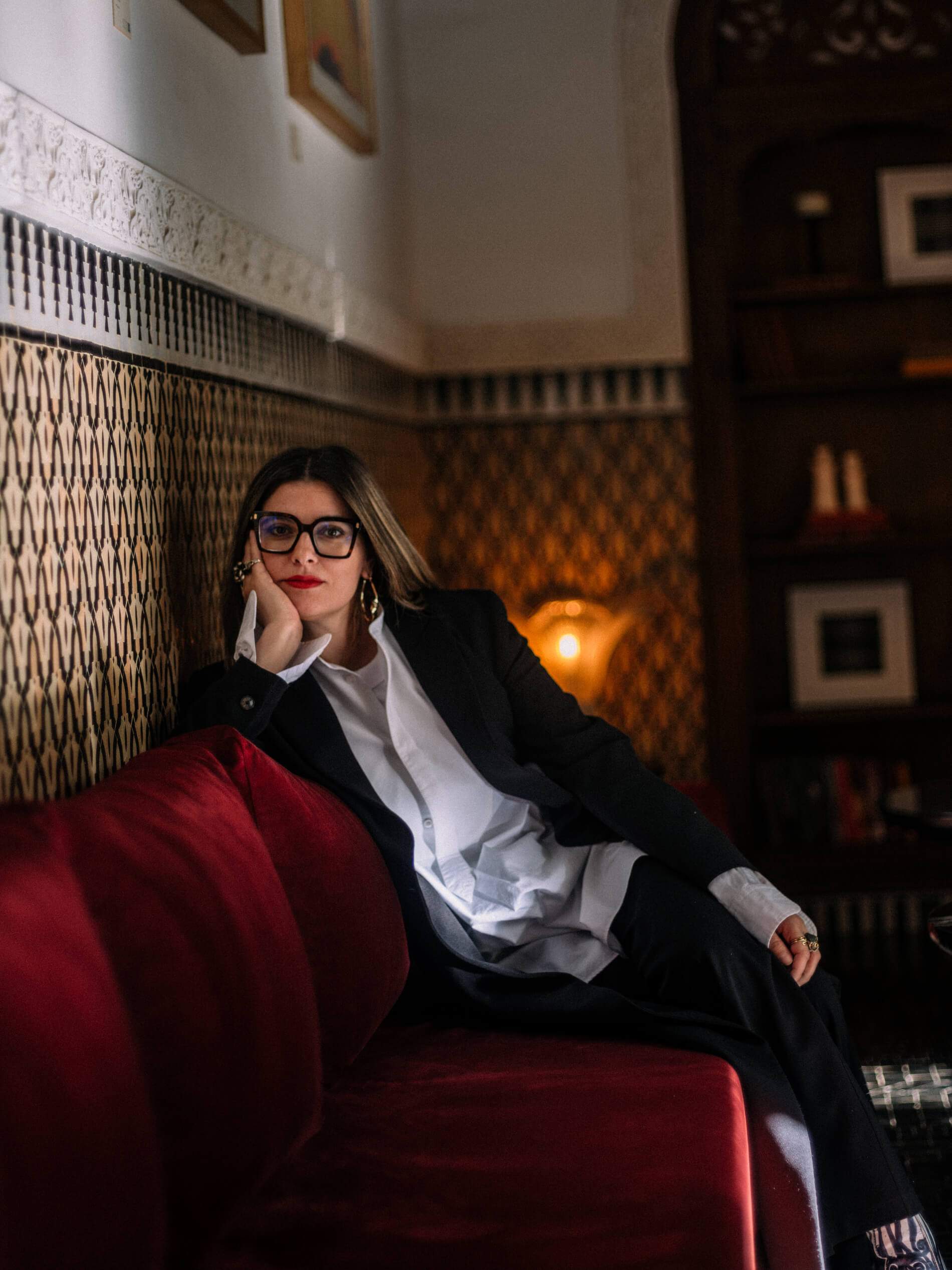 A woman wearing glasses and a black suit sits on a red couch.