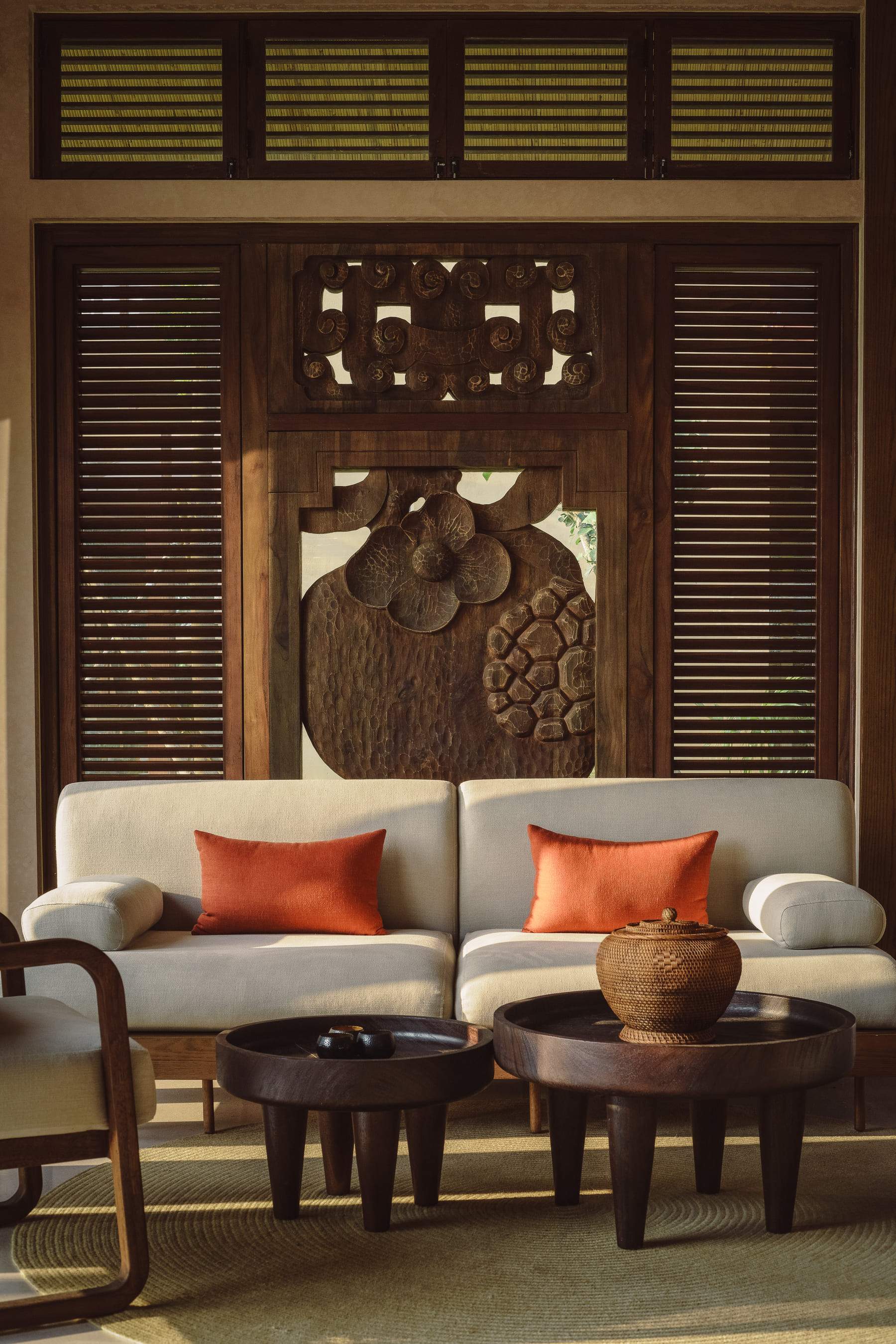 A white couch with two orange pillows and a wooden coffee table in front of a wooden wall with a carved design.