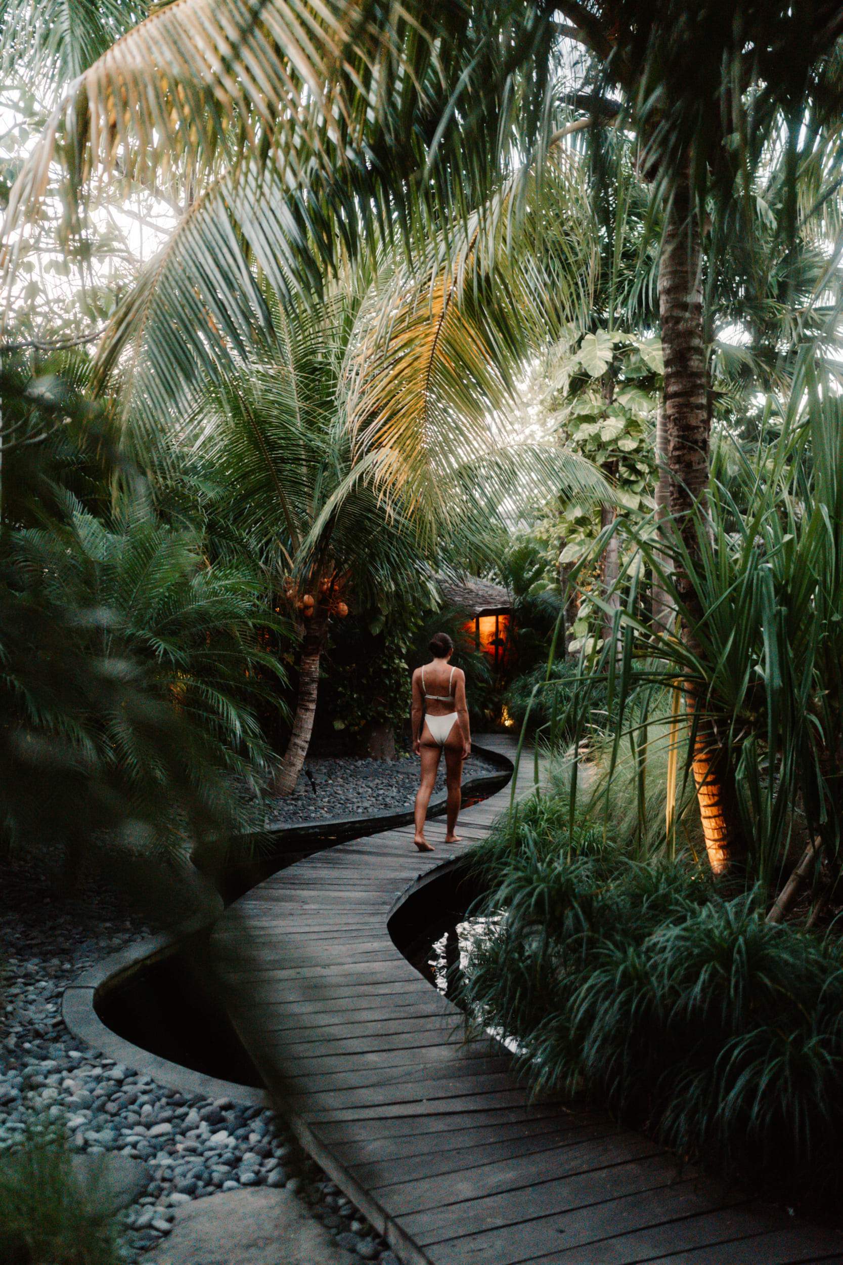 A woman wearing a white bikini walks on a wooden path in a jungle.