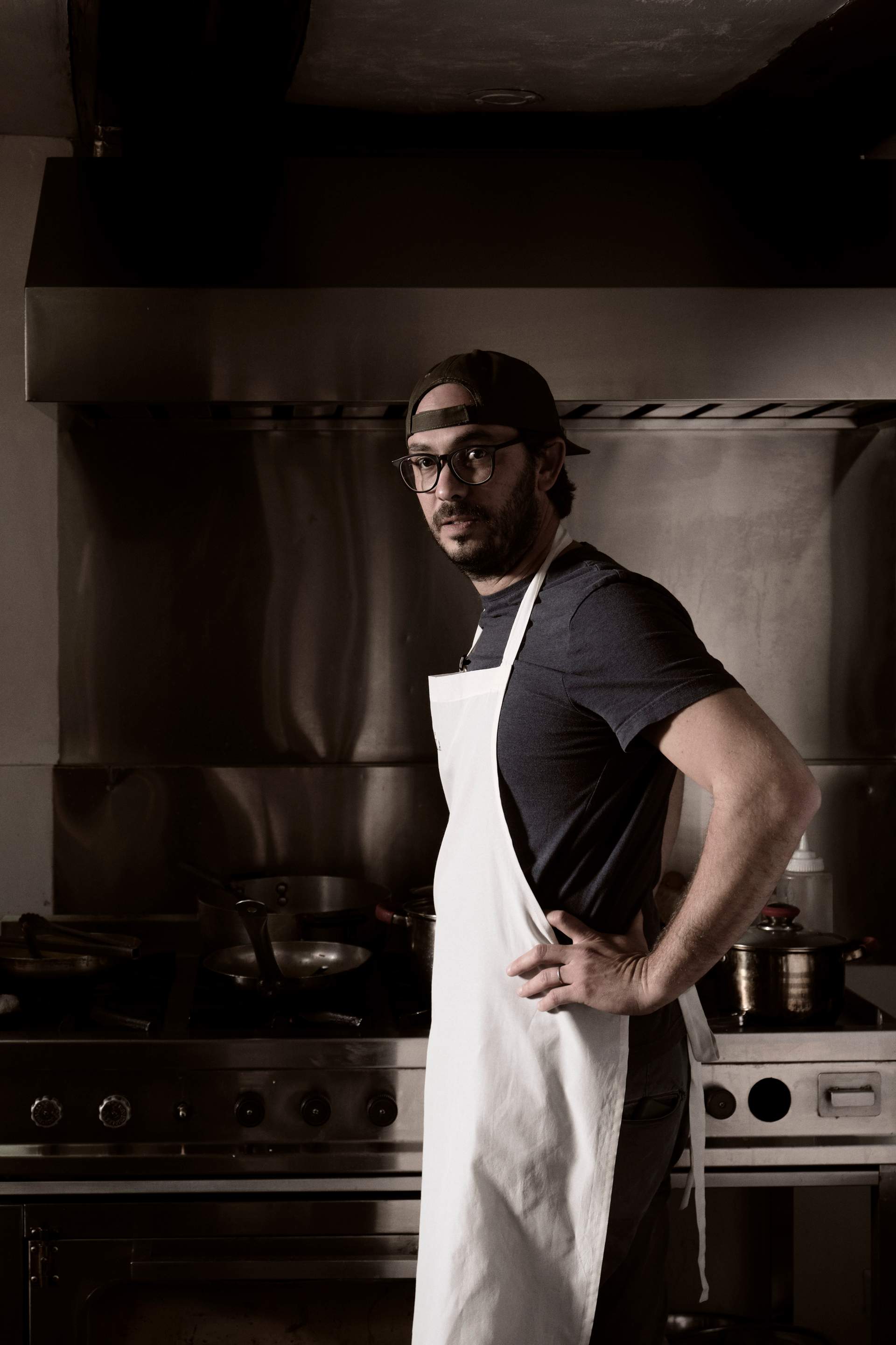 A man wearing glasses and a white apron stands in a kitchen.