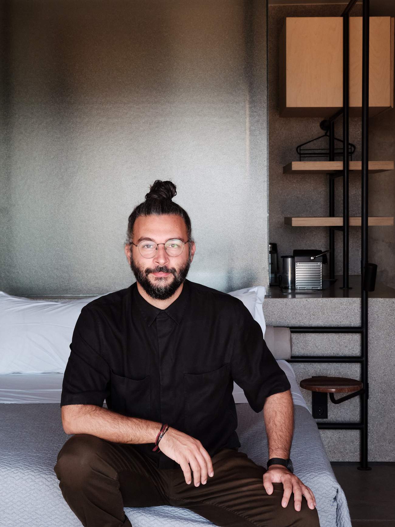 A man with a ponytail and glasses sitting on a bed.